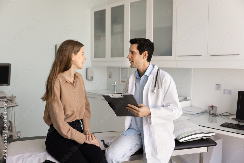Fototapeta premium Male doctor sits with female patient in clinic exam room, reviewing test results, explaining treatment options using clipboard, asking questions about symptoms. Communication, medical consultation