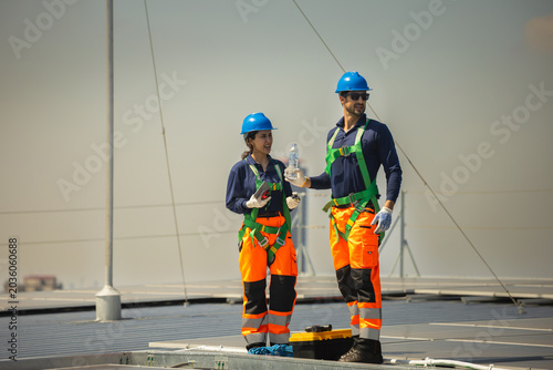 Maintenance team of technician and worker inspecting solar panels installation on industrial solar roof, Two solar engineers in safety uniform and harnesses working on rooftop solar farm
