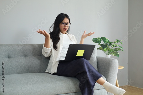 Tableau sur toile A young working woman sits on a sofa in the living room, staring anxiously at a laptop screen, reacting to bad and unexpected news, frustrated and angry, shrugging her shoulders and hands