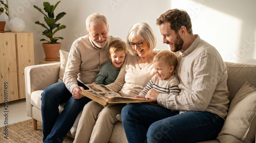Three generations share an old photo album on a sofa. Grandparents, adult son, and two grandchildren laugh together. Warm sunlight fills the Scandinavian living room.