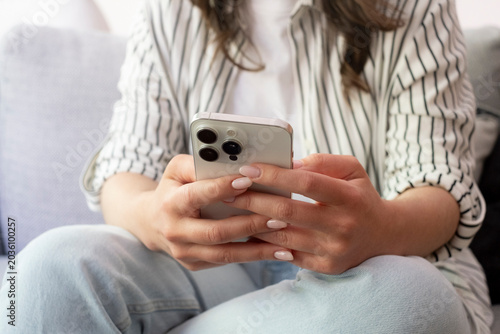 A relaxed woman sitting on a couch holding a smartphone in both hands, scrolling and texting while wearing a striped shirt and jeans, focused on social media, communication and digital lifestyle.