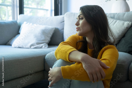 фотографія A pensive woman in a yellow sweater sits on the floor by a gray couch in a softl