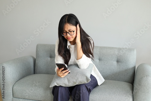 A young woman looks sad while covering her ears, sitting on the sofa at home, using her mobile phone, receiving bad news.