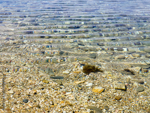 Clear Shallow Water Over Pebble Beach With Gentle Ripples And Sunlit Stones At Shoreline