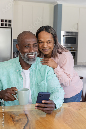 Diverse mature couple sitting at wooden kitchen table holding smartphone and ceramic mug