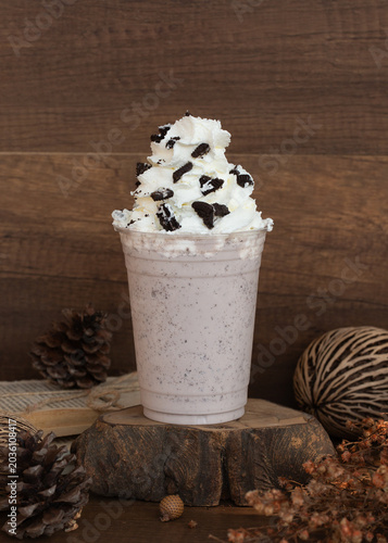 Cookies and cream milkshake smoothie in a plastic takeaway cup topped with whipped cream and crushed chocolate cookies on a rustic wood slice.