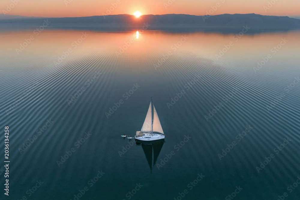 Fototapeta premium Calm sea surface with sailboat and paddleboarders at sunset for travel brochures, luxury lifestyle advertising and marine tourism background with distant mountains