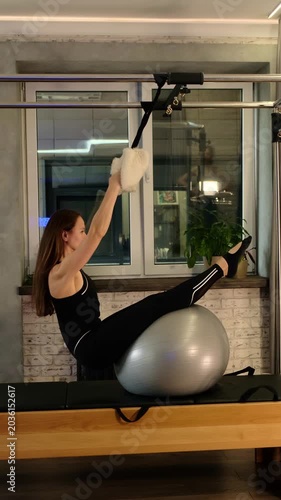 A woman reclines over a silver stability ball on Pilates Cadillac equipment, gripping soft overhead straps. The image reflects premium studio service, spinal mobility, body control, and calm wellness.