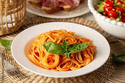 Classic Italian lunch with spaghetti al pomodoro and fresh basil. Traditional pasta with tomato sauce , with salad and bread with salumi in the background. Horizontal image.