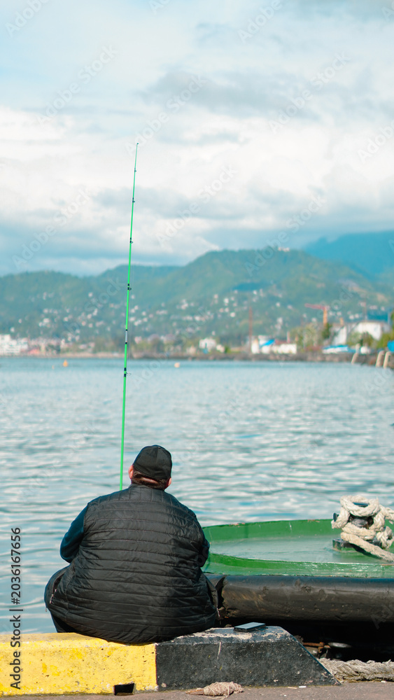 Obraz premium A man sits on a harbor pier fishing in calm waters with scenic mountains behind.