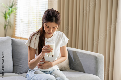 Fotografía Young asian woman sitting on grey sofa holding glass of milk touching stomach su