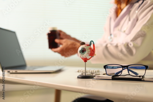 medical professional in a white coat writes patient records on a clipboard. An anatomical eye model, prescription pills, and a stethoscope