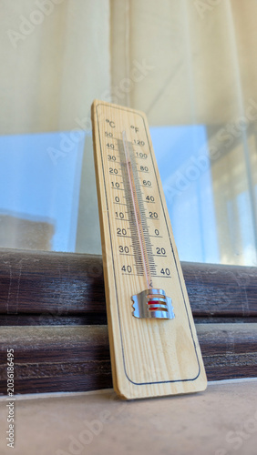 a wooden thermometer leaning on a window against blue sky and curtain representing abnormal summer heat wave extreme temperatures global warming ecological crisis and danger for people