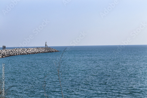 Rocky Breakwater and Calm Ocean Horizon Under Clear Sky. Stone Pier Extending Into Blue Sea with Small Lighthouse. Coastal Breakwater Structure in Deep Blue Ocean Water. Stone Jetty Distant Horizon
