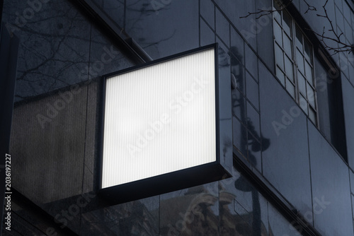 Blank illuminated square shop sign mockup on modern building exterior at dusk