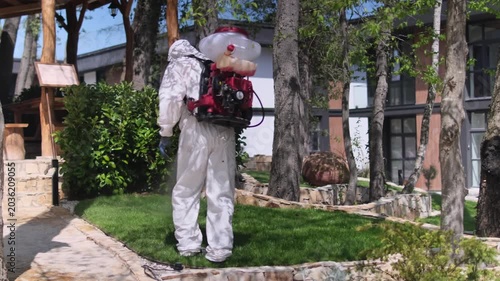 Worker in protective suit spraying garden with pesticide using backpack fogger, outdoor pest control treatment in residential yard