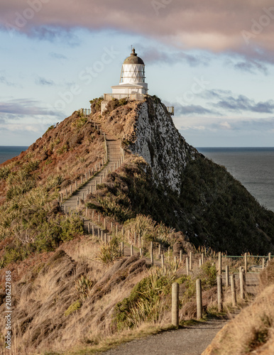 Lighthouse on coastal path under dramatic storm clouds - White coastal lighthouse viewed from a narrow path through windswept vegetation, with dark storm clouds creating a dramatic seascape atmosphere