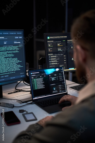 Bild auf Leinwand Vertical shot of young adult man monitoring cybersecurity dashboards across laptop and desktop screens, analyzing network data and threat alerts
