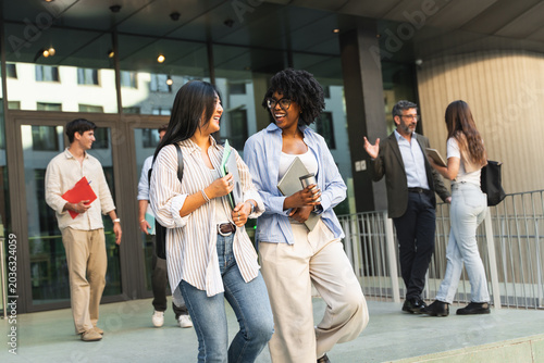 Two happy Asian African American students girls walking and talking in University campus after classes, going out of college