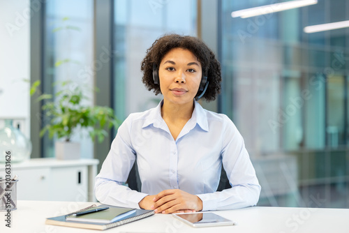 Portrait of a young African American woman wearing a headset sitting at an office desk and looking seriously at the camera