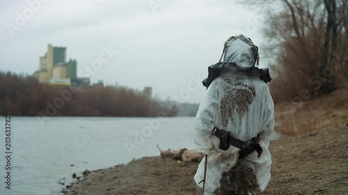 Field technician unknown in protective suit surveying toxic shore, respirator mask and heavy boots methodically sampling along river edge, distant factory skyline under gray sky, monitoring equipment