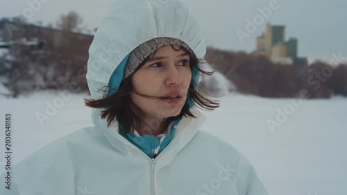 Asian scientist in white hazard suit standing on frozen river near abandoned plant, hood pulled up, breath visible, gaze scanning icy shoreline, expression tense and focused, field researcher
