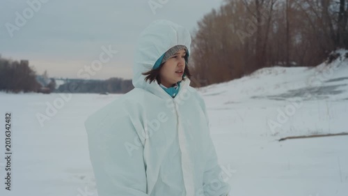 Woman in protective hooded suit walking along frozen shoreline, wide shot of lone figure scanning horizon, footprints in snow, overcast sky and distant tree line, steady reconnaissance with calm
