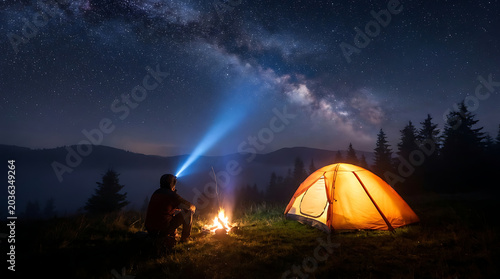 Person sitting near campfire under starry night sky with Milky Way and tent