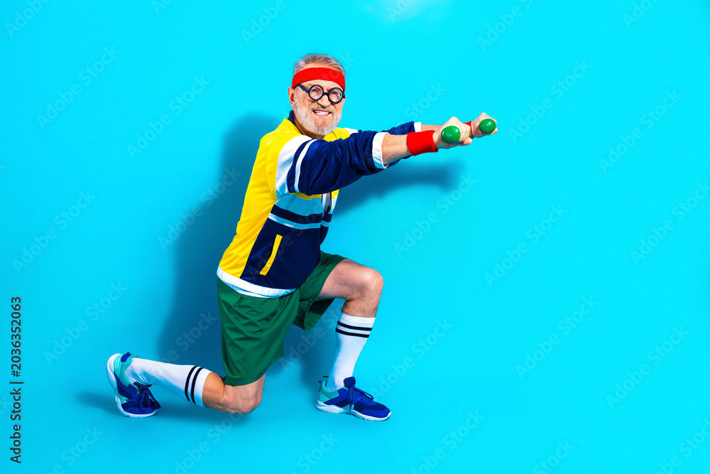 Obraz premium Senior man exercising with dumbbells in retro workout outfit on bright blue background, smiling and kneeling in a playful fitness pose promoting active lifestyle and joyful aging