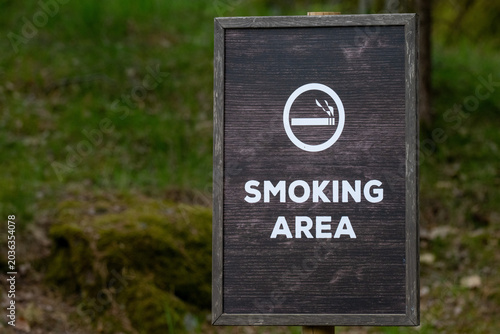 Slika na platnu A wooden sign stands in a park, showing a symbol and text that marks a designated smoking area