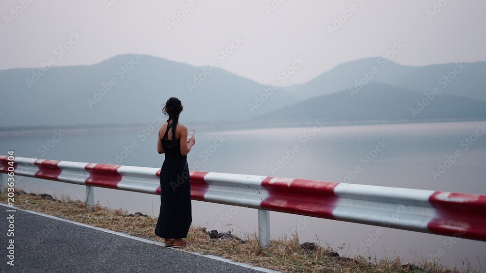Fototapeta premium Young woman standing on road observing tranquil lake with distant mountains