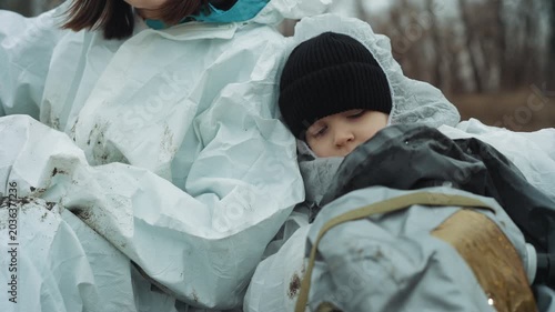 Child held in protective suit, field scientist cradling bundled child in white hazmat, beanie and backpack visible, mudstained fabric, barren winter woods background, tense tender mood, evacuation