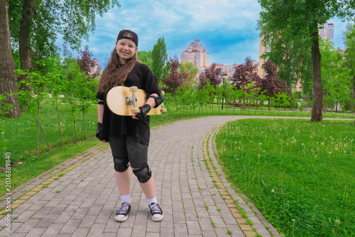 Happy young girl wearing black clothes and protective gear stands on a paved path in a green city park while holding her skateboard and smiling