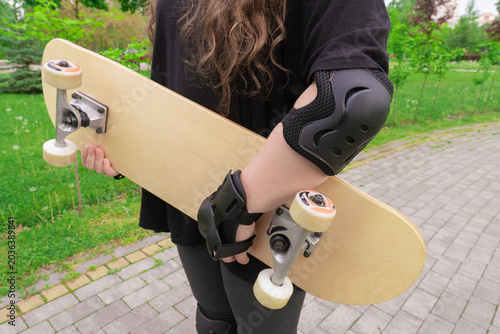 Close up of a young woman with long curly hair holding a blank wooden skateboard deck wearing protective gear in a summer park outdoors