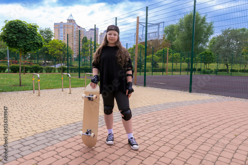 Teenage girl skater wearing knee pads, elbow pads, and wrist guards, standing with a blank wooden skateboard on a paved path in a city park