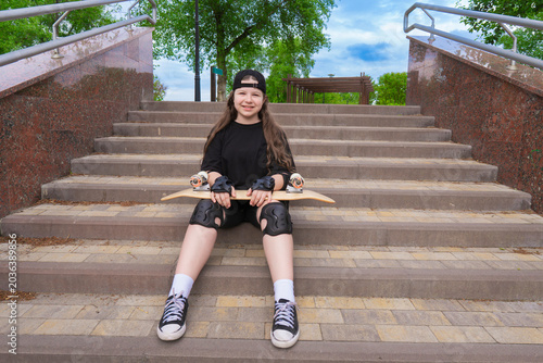 Happy teen girl with protective gear sitting on wide stone steps of city park holding skateboard on her knees and looking at camera