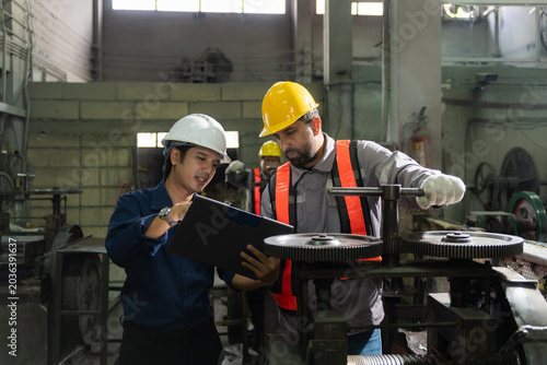 Professional manager with clipboard instructing and discussing maintenance plan with an industrial machinery operator turning gears inside a traditional factory manufacturing workshop