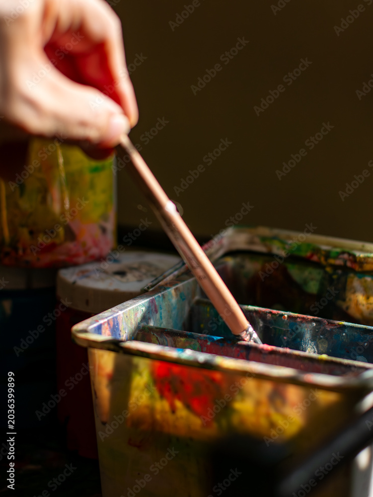 Obraz premium Artist hand stirring paint brush in colorful messy metal water container with acrylic paint jar and brush collection in art studio