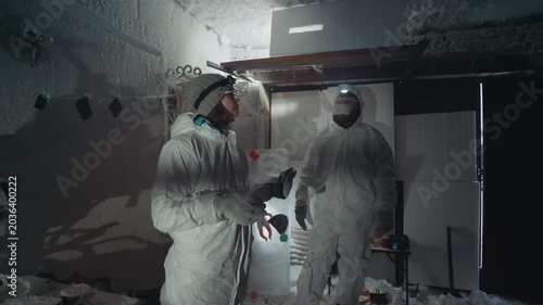 White technicians confer in contaminated shelter debriefing radiation readings beside doorway inspector compares dosimeter, medic secures sample, operator prepares containment during tense winter