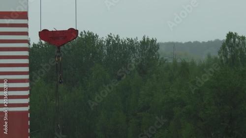 Close up view of heavy duty red crane hook and chain assembly, part of tall red and white striped crane mast, towering above dense green forest under flat gray overcast sky.