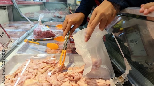 An unrecognisable woman uses a thong to transfer fresh chicken skin into a plastic bag, carefully packaging the meat for purchase at a market or supermarket.