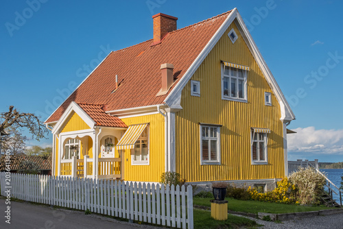 Принти на полотні Traditional yellow wooden house with red tiled roof and white picket fence by the lakeside in Sweden on a sunny spring day