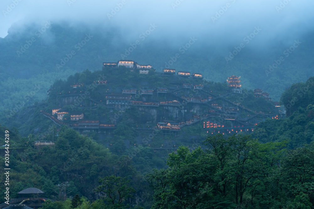 Fototapeta premium Traditional buildings with glowing lights stand on the green mountain slopes of Wangxian Valley Scenic Area in Jiangxi China during a foggy evening