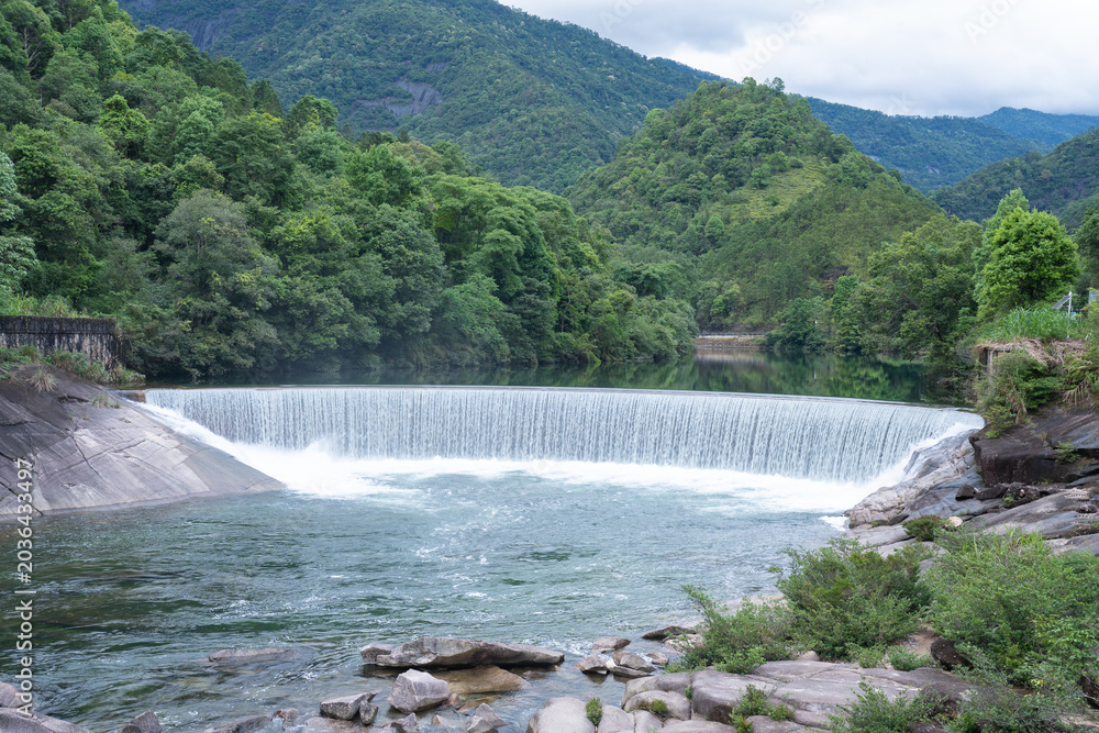 Obraz premium A scenic waterfall flows over a stone dam surrounded by lush green forests in the Wuyi Mountains of Fujian China