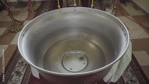 Metallic baptism font with clear water, on patterned rug inside church. Background shows candles, religious objects, indicating preparation for Christian baptism, sacrament, spiritual purification.
