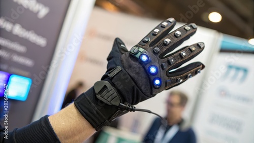 A futuristic black glove with embedded blue LED lights worn on a hand, showcasing advanced wearable technology in a blurred indoor setting.