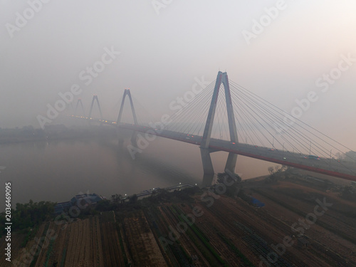 Morning fog surrounds a long cable stayed bridge crossing a calm river above agricultural fields in northern Vietnam.