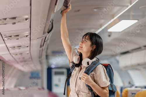 Young female traveler boarding an airplane, placing carry on luggage in overhead compartment, checking smartphone and passport while finding her seat before departure for her journey.