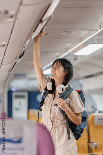 Young female traveler boarding an airplane, placing carry on luggage in overhead compartment, checking smartphone and passport while finding her seat before departure for her journey.