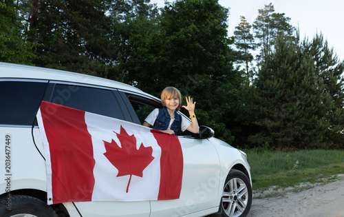 large Canadian flag hangs from the window of a white car, and a child looks at it and cheerfully waves his hand. Canada Day. National Identity. Travel around country. national symbol